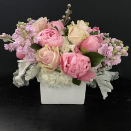 Low arrangement of pink peonies and blush roses in a white square vase with soft white flowers.