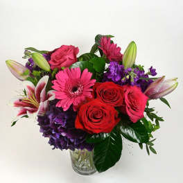 Mixed bouquet of roses, lilies, gerbera daisies, and purple hydrangea in a glass vase