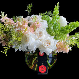 Low round arrangement of white roses and soft pink flowers in a clear bowl vase with airy greenery.