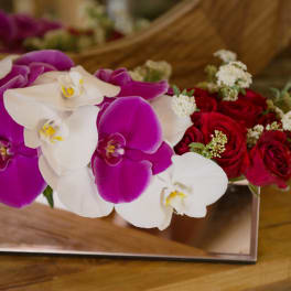 Purple and white orchids with red roses in a mirrored rectangular vase