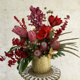 Red and purple floral arrangement in a gold vase on a woven mat