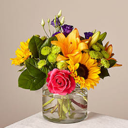 Mixed bouquet of sunflowers, roses, lilies, and purple blooms in a glass vase