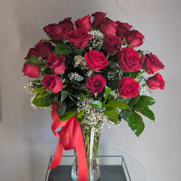 Tall arrangement of red roses with white filler flowers in a clear vase with a red ribbon bow