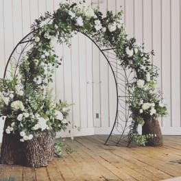 White floral arch with greenery on a black metal frame and tree-stump bases