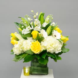 Yellow and white floral arrangement in a glass vase with a small candle beside it.