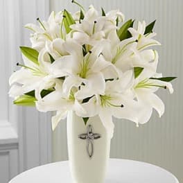 White lilies arranged in a tall cream vase with a silver cross emblem