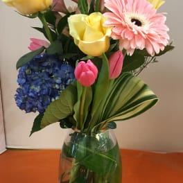 Mixed bouquet in a clear glass vase with yellow roses, pink gerbera, blue hydrangea, and tulips