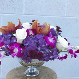 Purple hydrangeas, white roses, and orchids in a silver pedestal bowl
