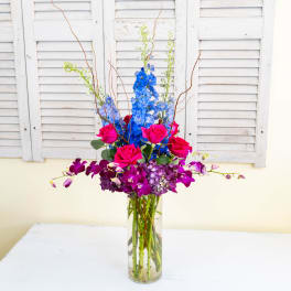 Tall vase arrangement with blue flowers, pink roses, and purple orchids