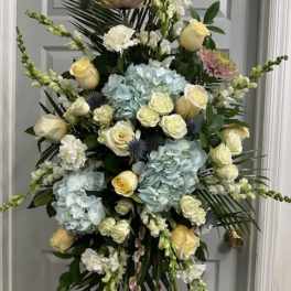 Tall funeral spray with pale roses, blue hydrangeas, and white blooms on a stand