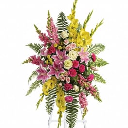 Tall standing spray with pink lilies, roses, carnations, yellow blooms, and ferns on a white easel.