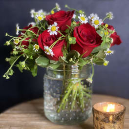 Red roses and white daisies in a glass jar beside a lit candle