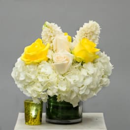 White hydrangeas and yellow roses in a glass vase with a gold candle holder beside it