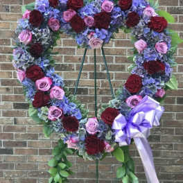 Heart-shaped standing wreath of red and pink roses with purple hydrangeas and trailing greenery on an easel.