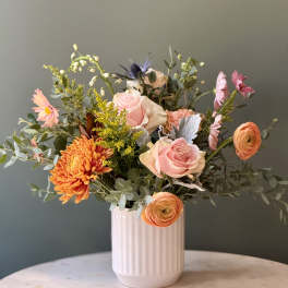 Mixed arrangement of peach roses, orange mums, and ranunculus in a white ribbed ceramic vase