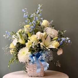 White, yellow, and blue flower basket with lilies, roses, mums, and a blue ribbon bow