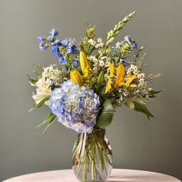 Mixed bouquet with blue hydrangea, yellow lilies, and white daisies in a clear glass vase.