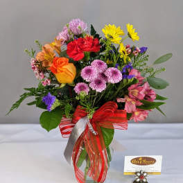 Bright mixed bouquet of roses, carnations, daisies, and mums in a clear vase with a red bow.