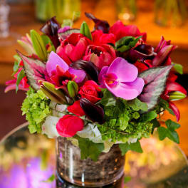 Compact arrangement of pink orchids, red blooms, and green accents in a glass vase on a mirror