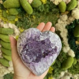 Hand holding a heart-shaped purple crystal against a green floral wall
