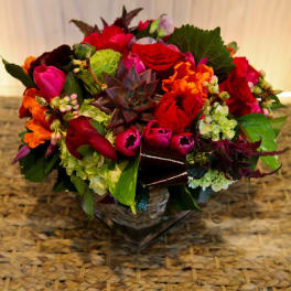 Low mixed arrangement of red roses, pink tulips, green hydrangeas, and a central succulent in a glass cube vase.