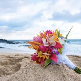 Bright tropical hand-tied bouquet with pink, orange, and yellow flowers resting on sand by the ocean