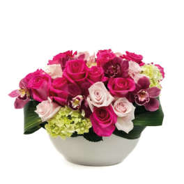 Low arrangement of hot pink and pale pink roses with orchids and hydrangea in a white bowl.