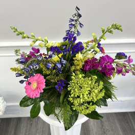 Bright mixed arrangement with pink gerbera, purple flowers, and yellow snapdragons displayed on a white pedestal.