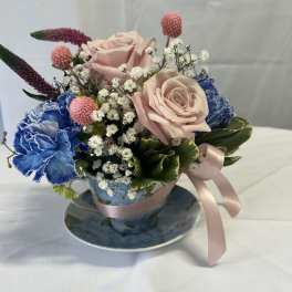 Pink roses and blue carnations arranged in a teacup with a ribbon