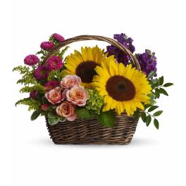 Basket arrangement with yellow sunflowers, pink roses, and purple flowers in a woven handled basket.