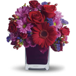 Compact arrangement of red roses, red gerbera daisy, and purple mums in a clear square glass vase