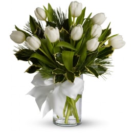White tulips with holiday evergreens in a clear glass vase tied with a white bow.
