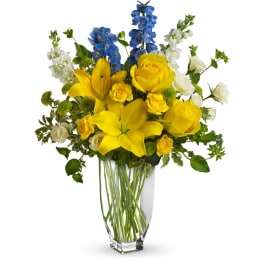 Tall glass vase with yellow lilies, yellow and white roses, and blue delphinium.