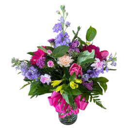 Mixed bouquet of pink, purple, yellow, and peach flowers in a clear glass vase with a bright pink ribbon bow