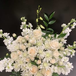 Cream roses and white flowers arranged in a low bouquet against a dark background.
