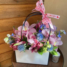 Pink orchid arrangement in a white gift bag with a heart ribbon