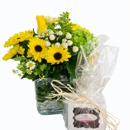 Yellow flower arrangement in a glass cube vase with a wrapped gift box beside it