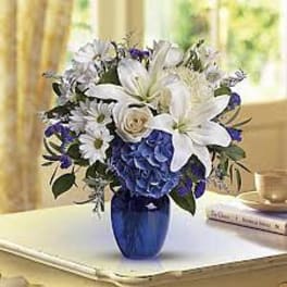 White lilies and daisies with blue hydrangeas in a cobalt blue glass vase on a table
