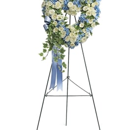 Heart-shaped blue and white floral wreath with ivy and blue ribbon on a tall easel stand.