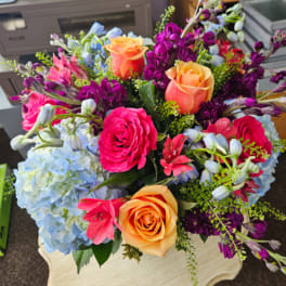 Bright bouquet with orange and pink roses, blue hydrangeas, and purple flowers on a small pedestal table