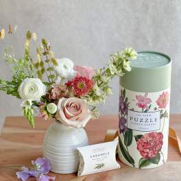 Mixed flower arrangement in a white vase beside a floral puzzle tin and caramel box