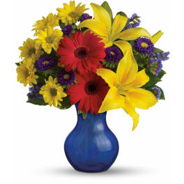Arrangement of yellow lilies, red gerberas, and mixed blooms in a blue glass vase