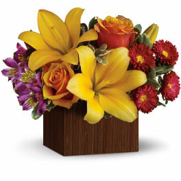 Compact arrangement of yellow lilies, orange roses, and red and purple blooms in a brown cube vase