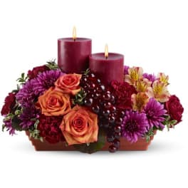 Low centerpiece of orange roses, burgundy carnations, purple mums and two burgundy pillar candles in a rectangular container