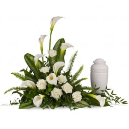 White calla lilies and roses arranged around greenery beside a white urn.