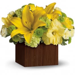 Low arrangement of yellow lilies and mixed green and white flowers in a dark wooden cube vase