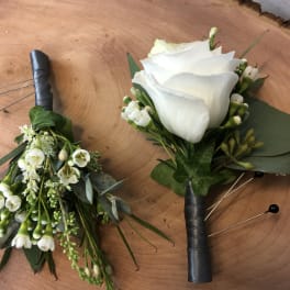 Two white floral boutonnieres with black wrapped stems on a wood surface.
