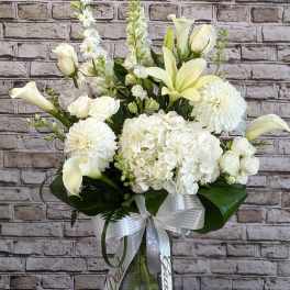 Tall white floral arrangement with lilies, roses, hydrangeas and calla lilies in a glass vase with white ribbon