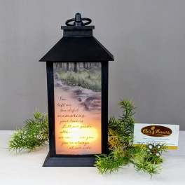 Black memorial lantern with glowing light and verse, accented by small green sprigs.