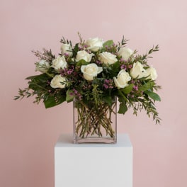 Arrangement of white roses with pink filler flowers in a clear glass cube vase against a pink background